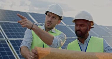 Erkek mühendislerin güneş enerjisi çiftliğinde dikilirken iletişim ve kontrol planlarını yakından inceleyin. Üniformalı ve kasklı adamlar projeyi tartışıyor ve geleceğe bakıyor..