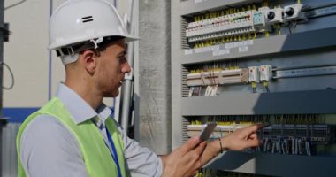 Güneş panelinin terminal kutusunu kontrol ederken yakışıklı erkek mühendisin tablet kullandığı görüntüyü kapat. Kasklı ve üniformalı bir adam elinde aletle kontrol panelinin yanında duruyor..