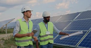 Tabletli ve parşömenli erkek mühendisler iletişim kuruyor. Üniformalı ve kasklı iki adam güneş çiftliğinde yürürken projeyi tartışıyorlar. Yeşil enerji kavramı.