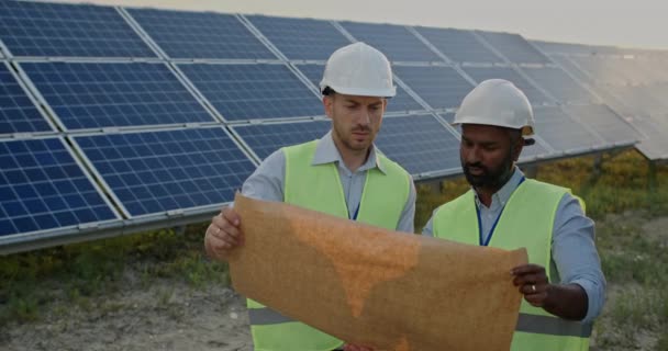 太陽光発電所に立つ2人の男性技術者のミステリービューと建設の効率的な計画を議論する。ハードヘルメットと均一な話とエンジニアリング図面を見て男性. — ストック動画