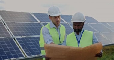 Melez ırk erkek mühendislerinin güneş panellerindeki sıra sıra dizilişi ve verimli bir inşaat planı tartışmaları. Kasklı ve üniformalı adamlar iletişim kuruyor ve planlara bakıyor..