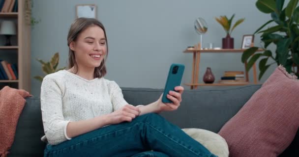 Dove duizendjarige vrouw met videogesprek en tonen met gebarentaal zin Goed nieuws terwijl zittend op de bank. Vrouwelijke persoon glimlachend tijdens het gebruik van smartphone voor communicatie. Gehoorverlies. — Stockvideo