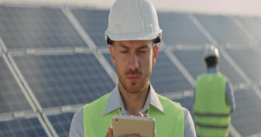 Erkek mühendisin tablet ekrana baktığı görüntüyü kapatın. Sert kasklı ve üniformalı bir adam yenilenebilir enerji çiftliğinde duruyor ve alet kullanıyor. Arka plandaki güneş panelleri. Anamorfik lens.