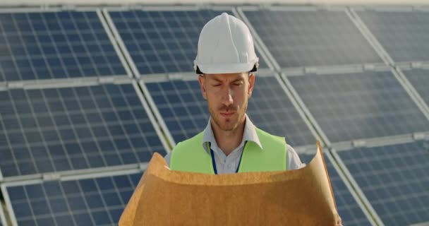 Retrato de belo trabalhador masculino olhando para o rolo de desenho de engenharia e do que para a câmera. Homem de capacete duro em pé na fazenda de energias renováveis. Painéis solares no fundo. Lente anamórfica. — Vídeo de Stock