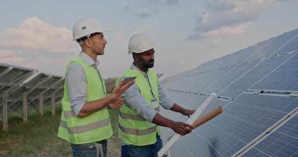 Engenheiros de corrida mistos com tablet e pergaminhos de engenharia de desenho conversando. Homens de capacetes uniformes e duros discutindo projeto enquanto estão perto de painéis solares. Conceito de energia verde. — Vídeo de Stock