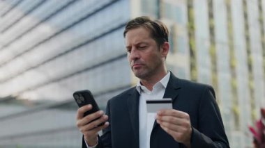 Odaklanmış girişimci portresi parayı akıllı telefon ve kredi kartıyla şehir caddesinde aktarıyor. Başarılı iş adamı ofis binasının yanında duran ödeme için bankacılık uygulaması kullanıyor