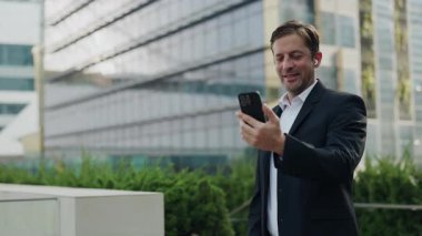 Ofis binasının dışındaki akıllı telefondan iş arkadaşlarına el sallayan dost canlısı iş adamının portresi. Kulaklıklı, olgun şirket CEO 'su sokakta online toplantıya katılıyor.