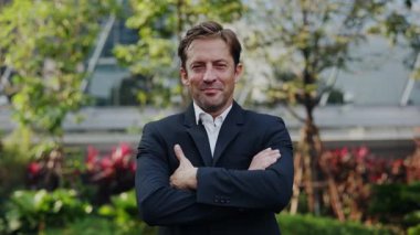 Takım elbiseli mutlu iş adamının portresi şehir parkında kollarını kavuşturuyor. Olgun girişimci, şehir merkezinde yavaş çekimde başarı ve profesyonelliği ifade ederken gülümsüyor.