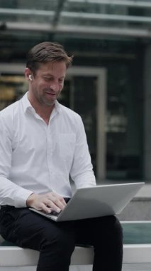 Dost canlısı iş adamının portresi ofis dışında bilgisayarla iş arkadaşlarıyla görüntülü görüşme yapıyor. Olgun takım lideri, kentin dikey çekimlerinde iş arkadaşlarını jest yaparak karşıladı