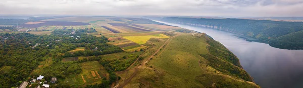 Panorama of big river canyon, village and rural fields on hills. Aerial view on green and yellow parts of fields in autumn season. River Dniester, Podolia, Ukraine