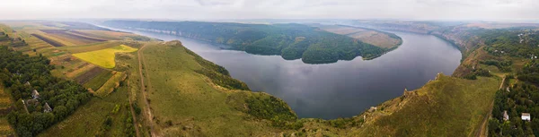 Panorama of big river canyon, village and rural fields on hills. Aerial view on green and yellow parts of fields in autumn season. River Dniester, Podolia, Ukraine