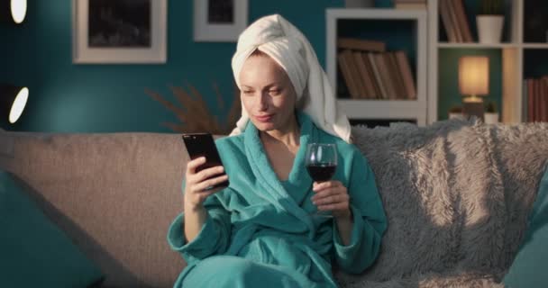Расслабленная женщина в халате с помощью телефона и питьевого вина — стоковое видео