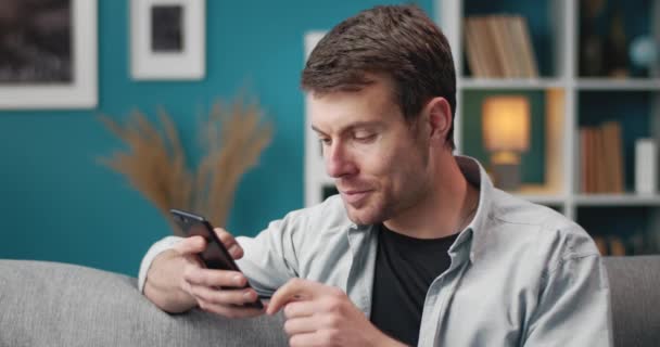 Sorrindo homem usando smartphone enquanto sentado no sofá — Vídeo de Stock