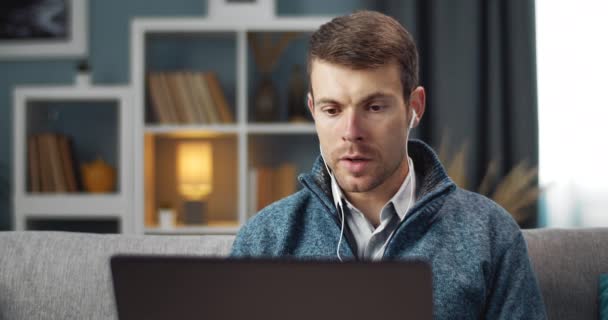 Man in earphones using laptop for video chat on couch — Stock videók