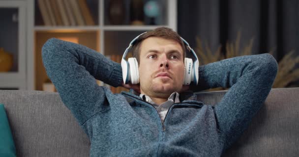 Young man relaxing on grey couch in wireless headphones — Stockvideo