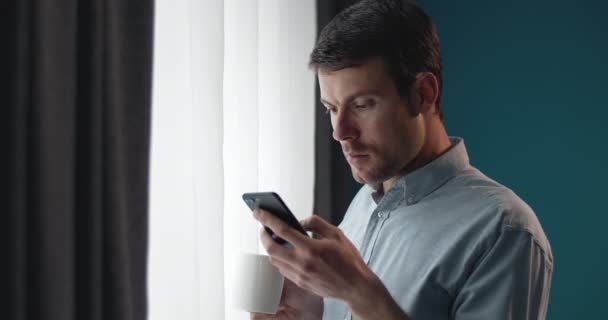 Hombre de pie cerca de la ventana con teléfono y taza de café — Vídeo de stock