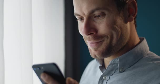 Chico feliz usando smartphone mientras está de pie cerca de la ventana — Vídeo de stock