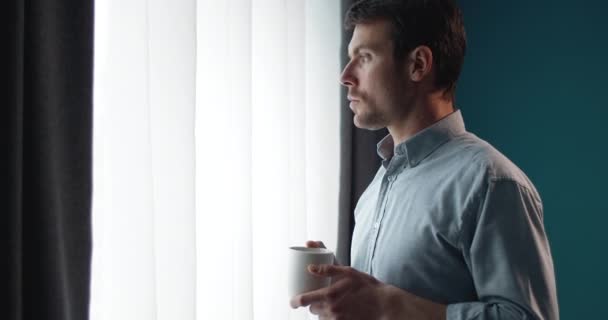 Красивый мужчина с чашкой кофе, выглядывающий в окно — стоковое видео
