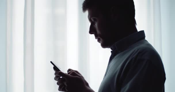 Silueta del hombre feliz utilizando el teléfono inteligente cerca de la ventana — Vídeos de Stock