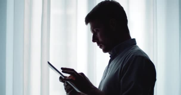 Smiling man in silhouette using digital tablet near window — Αρχείο Βίντεο