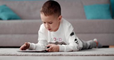 Mutlu çocuk halıda uzanıyor ve tablette oyunlar oynuyor.