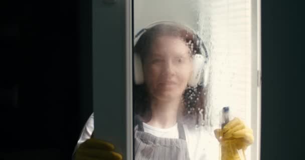 Mujer sonriente en auriculares limpiando vidrio de ventana en casa — Vídeo de stock