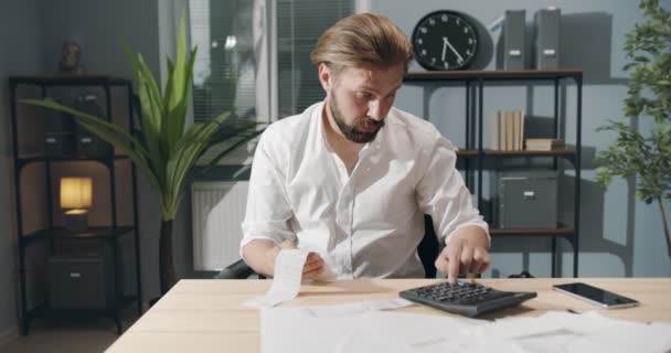 Vermoeide man met baard aan tafel die rekeningen berekent — Stockvideo