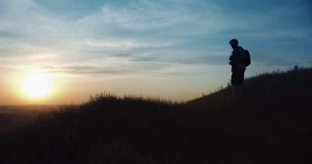 Человек в силуэте стоит на холме и делает фото заката — стоковое видео