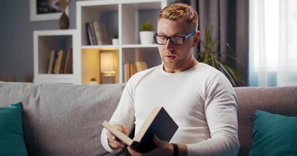 Knappe man voelt zich moe van het lezen van boek thuis — Stockvideo