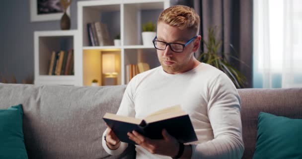 Jovem de óculos lendo livro enquanto sentado no sofá — Vídeo de Stock