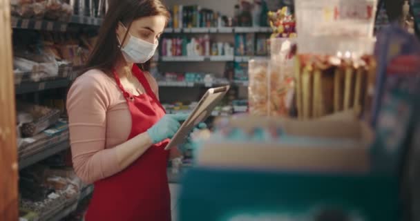 Chica en máscara y guantes haciendo revisión con la tableta en la tienda — Vídeos de Stock