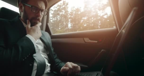 Στοχαστικός άνθρωπος που χρησιμοποιεί φορητό υπολογιστή, ενώ κάθεται στο πίσω κάθισμα του αυτοκινήτου — Αρχείο Βίντεο