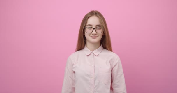 Chica en gafas y camisa rosa sonriendo y mirando a la cámara — Vídeos de Stock