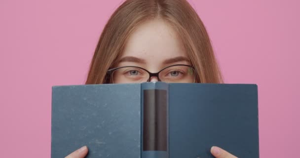 Mujer escondiendo la cara detrás de libro abierto sobre fondo rosa — Vídeos de Stock