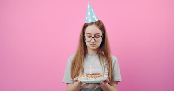 Upset girl in festive cap holding cake with candle in studio — Stock Video