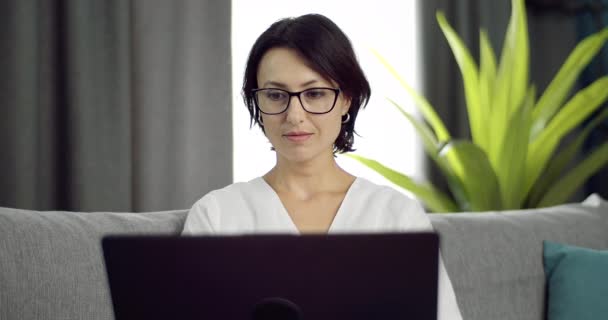 Θετική γυναίκα με γυαλιά που κάθεται στον καναπέ και χρησιμοποιεί φορητό υπολογιστή — Αρχείο Βίντεο