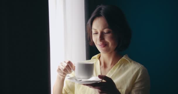 Vacker kvinna står nära fönstret och dricker kaffe — Stockvideo