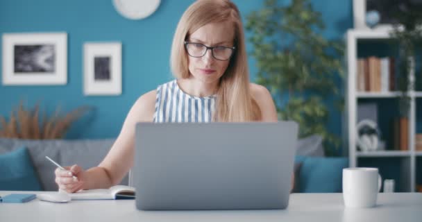 Mujer sentada a la mesa con portátil abierto y tomando notas — Vídeo de stock