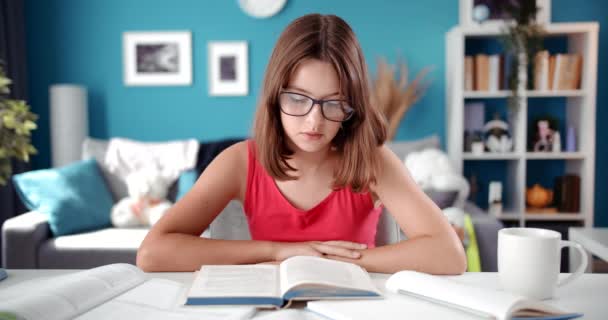Schulmädchen mit Brille sitzt am Tisch und liest Buch — Stockvideo
