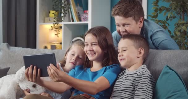 Quatro crianças olhando para a tela do tablet e sorrindo no sofá — Vídeo de Stock
