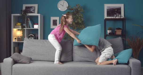 Χαμογελώντας μικρά παιδιά αγωνίζονται με μπλε μαξιλάρια σε γκρι καναπέ — Αρχείο Βίντεο