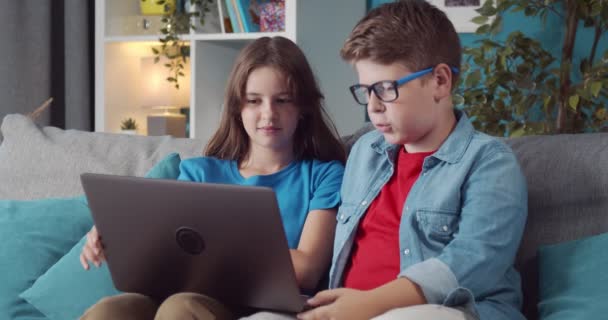 Två barn sitter på grå soffa och använder trådlös bärbar dator — Stockvideo