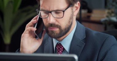 Cep telefonundan konuşan ve bilgisayar ekranına bakan sakallı adam.