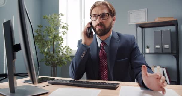 Mannen sitter vid bordet med datorn och pratar i mobilen — Stockvideo