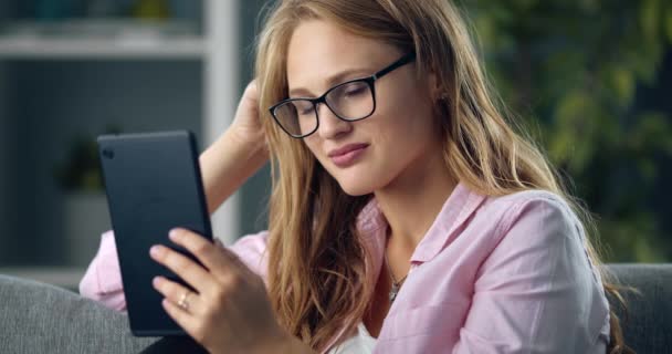 Ελκυστικό κορίτσι με ξανθά μαλλιά χρησιμοποιώντας ψηφιακό tablet στον καναπέ — Αρχείο Βίντεο