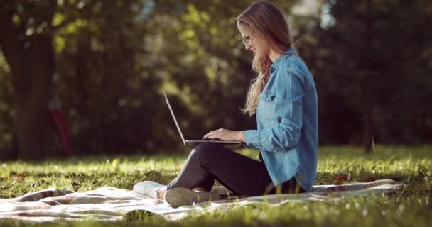Веселая юная леди печатает на ноутбуке, сидя в парке — стоковое видео