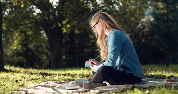 Γυναίκα κρατώντας σημειώσεις, ενώ σπουδάζουν σε φορητό υπολογιστή στο πάρκο — Αρχείο Βίντεο