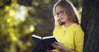 Güzel bir kadın parktaki ağacın yanında oturmuş kitap okuyor.