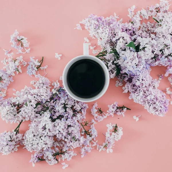 Flat lay cup of coffee with branches of lilac flowers on pink background. Top view spring minimal concept