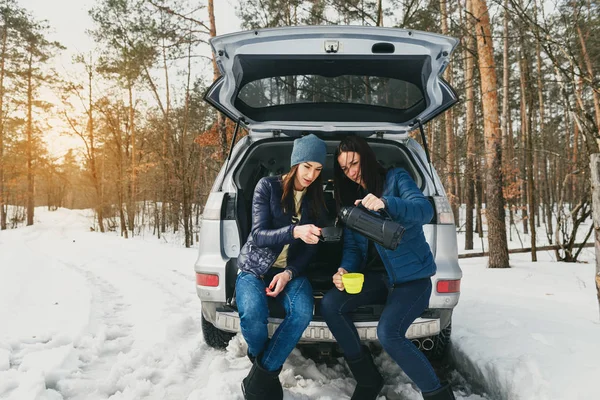 Two young women in winter wear on snowy day in the winter forest drinking coffee in the car